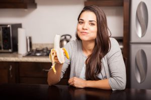 Cute young Hispanic woman chewing a banana while sitting in the kitchen