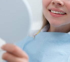 Close-up of a patient in a dentist's office examining her teeth by smiling and looking in a hand mirror.