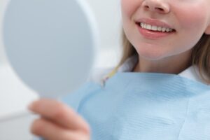 Close-up of a patient in a dentist's office examining her teeth by smiling and looking in a hand mirror.